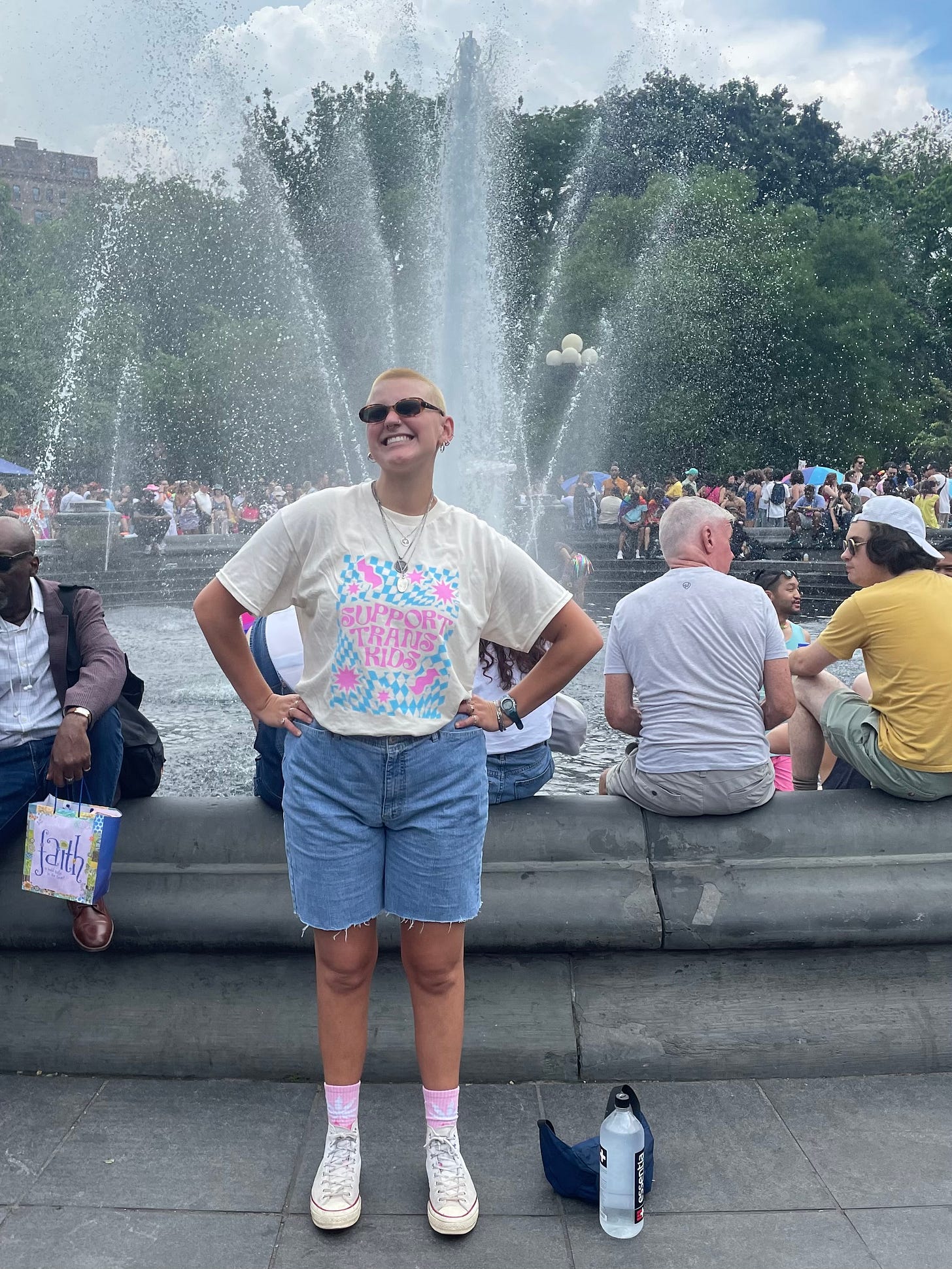 Myself at New York pride in 2023. I am standing in front of a fountain with my hands on my waist, a huge smile on my face. My head is shaved and bleached and I am wearing a shirt that reads “support trans kids.”