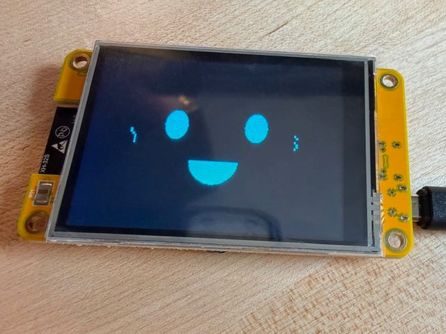Photo of a yellow ESP32 touchscreen on a wooden surface displaying a bright blue happy face with two eyes and a wide smile on a dark screen.