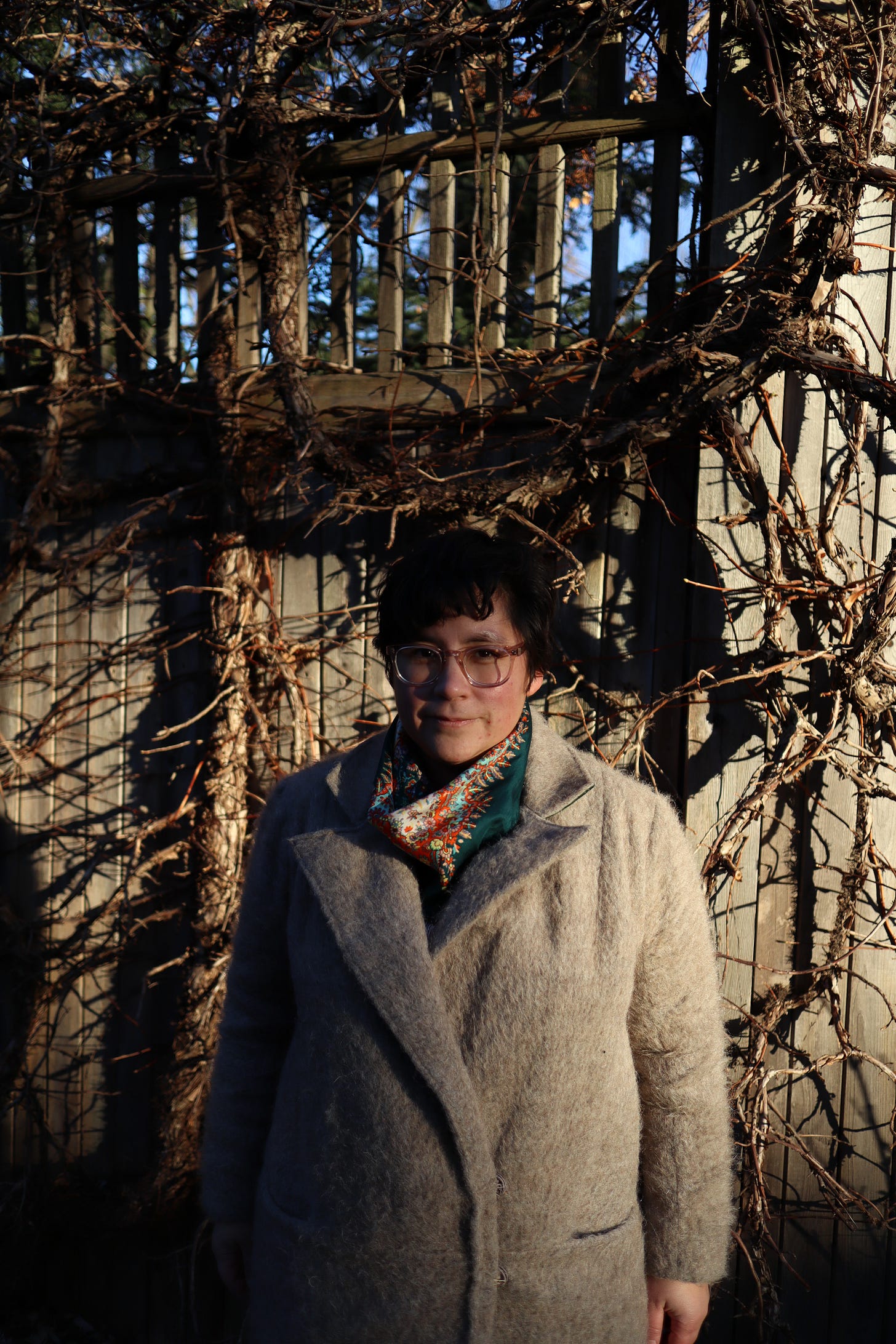 Christine standing in front of gnarly branches wearing her hand-sewn beige fuzzy coat with dark green scarf and pale glasses