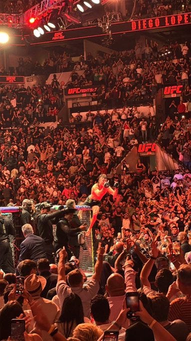Diego Lopes standing in a crowded arena, raising his arms in celebration. He is surrounded by people taking photos and cheering. UFC branding and lights are visible in the background, with a large screen displaying "Diego Lopes". Diego Lopes standing in a crowded arena, raising his arms in celebration. He is surrounded by people taking photos and cheering. UFC branding and lights are visible in the background, with a large screen displaying "Diego Lopes".