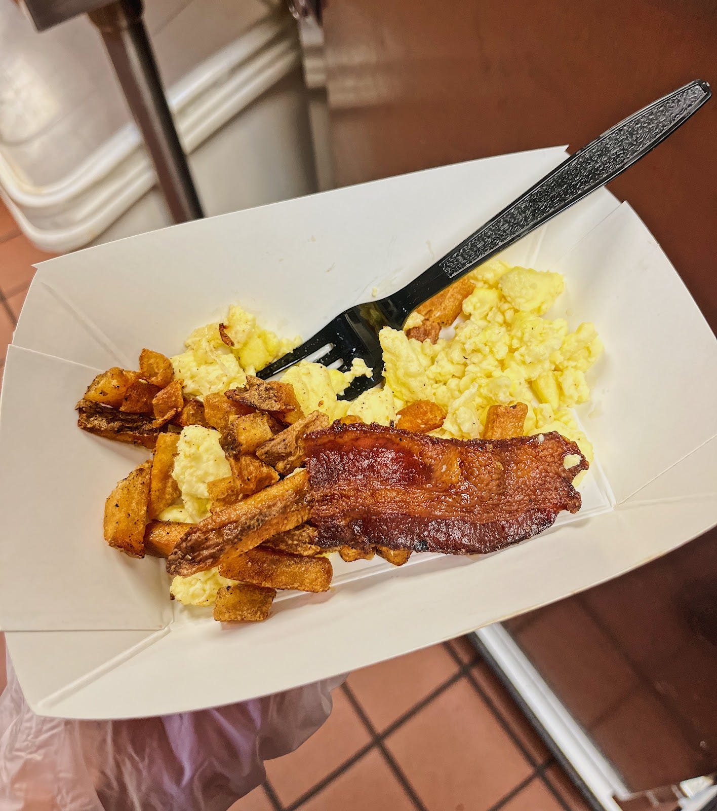 Scrambled eggs, a slice of bacon, and cut up french fries in a paper bowl in a food service gloved hand