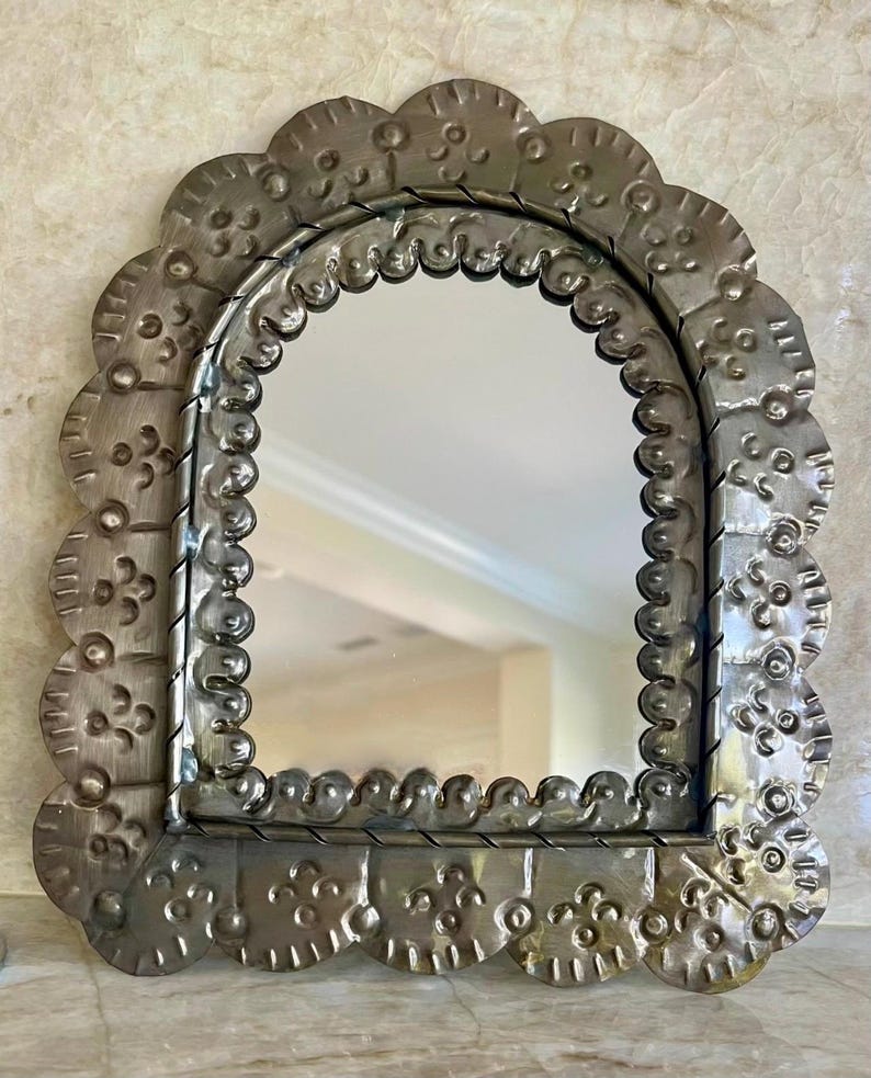 May include: A silver-toned, arched mirror with a decorative, scalloped frame. The frame is embossed with a floral pattern.