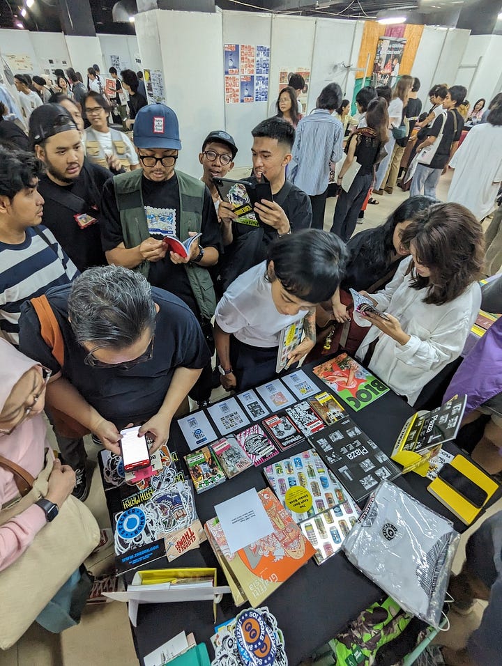 The first image is alarge outdoor banner advertising the Jakarta Art Book Fair 2023. The banner displays details such as the dates (27-29 October 2023), location (Pasaraya Blok M, Lower Ground), and hours (11 AM - 7 PM). The banner is surrounded by greenery with a tall white building visible in the background. The second image A busy scene at a booth during the Jakarta Art Book Fair 2023, with a group of people gathered around a table displaying books and stickers. Several attendees are browsing through the books and taking photos, while the table is covered with colorful books, sticker packs, and promotional materials.