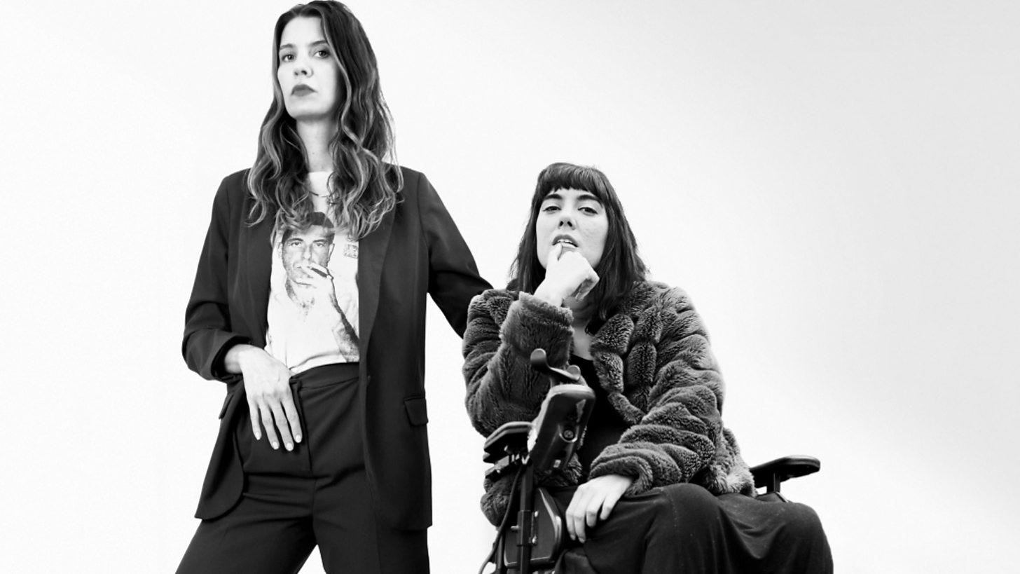 Two white women are in a black and white photo. One of them uses a powerchair, wearing a fleece coat.