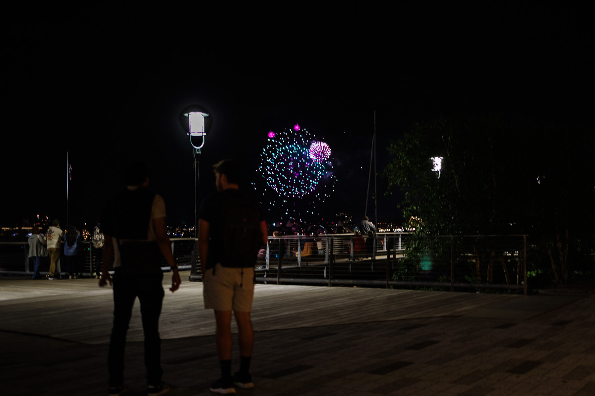 Two people standing on Seaport pier watching colorful fireworks light up Boston Harbor night sky