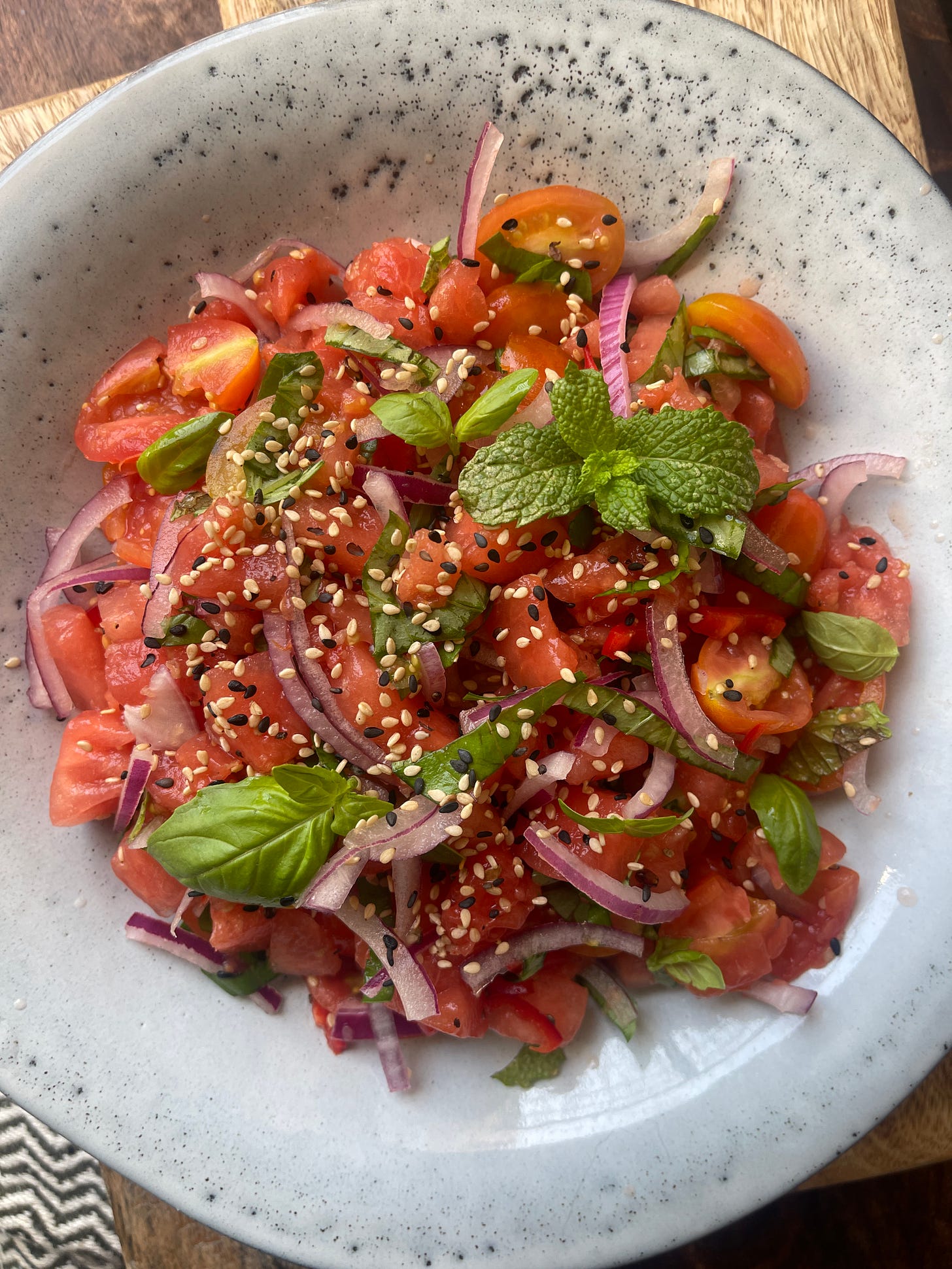 Overhead view of a Mediterranean watermelon salad with cherry tomatoes, red onion, fresh herbs, and sesame seeds, served in a handmade ceramic bowl on a wooden table