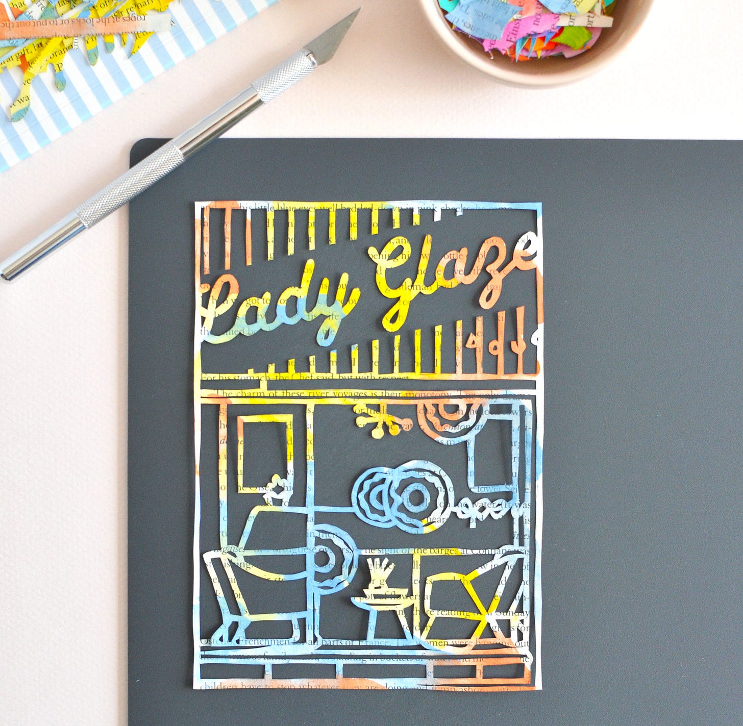 A colourful paper cutting depicting the storefront of a donut shop called Lady Glaze. Shown on a dark grey cutting mat with exacto knife and a little bowl filled with paper scraps.