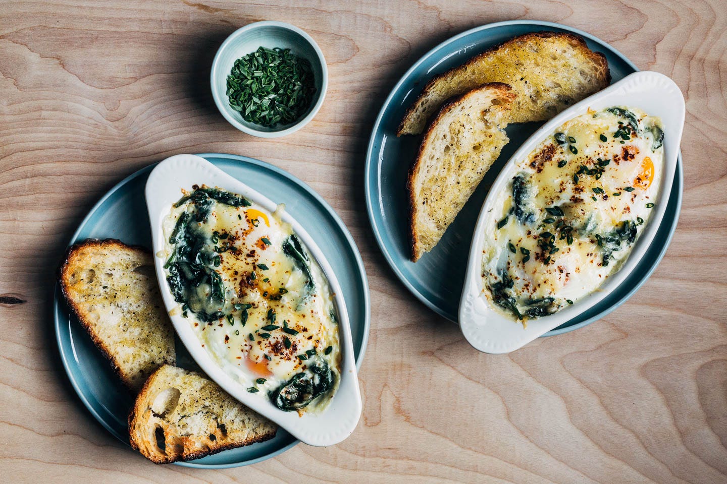 Two ramekins with baked eggs on plates with croutons, and a little dish with minced chives alongside.