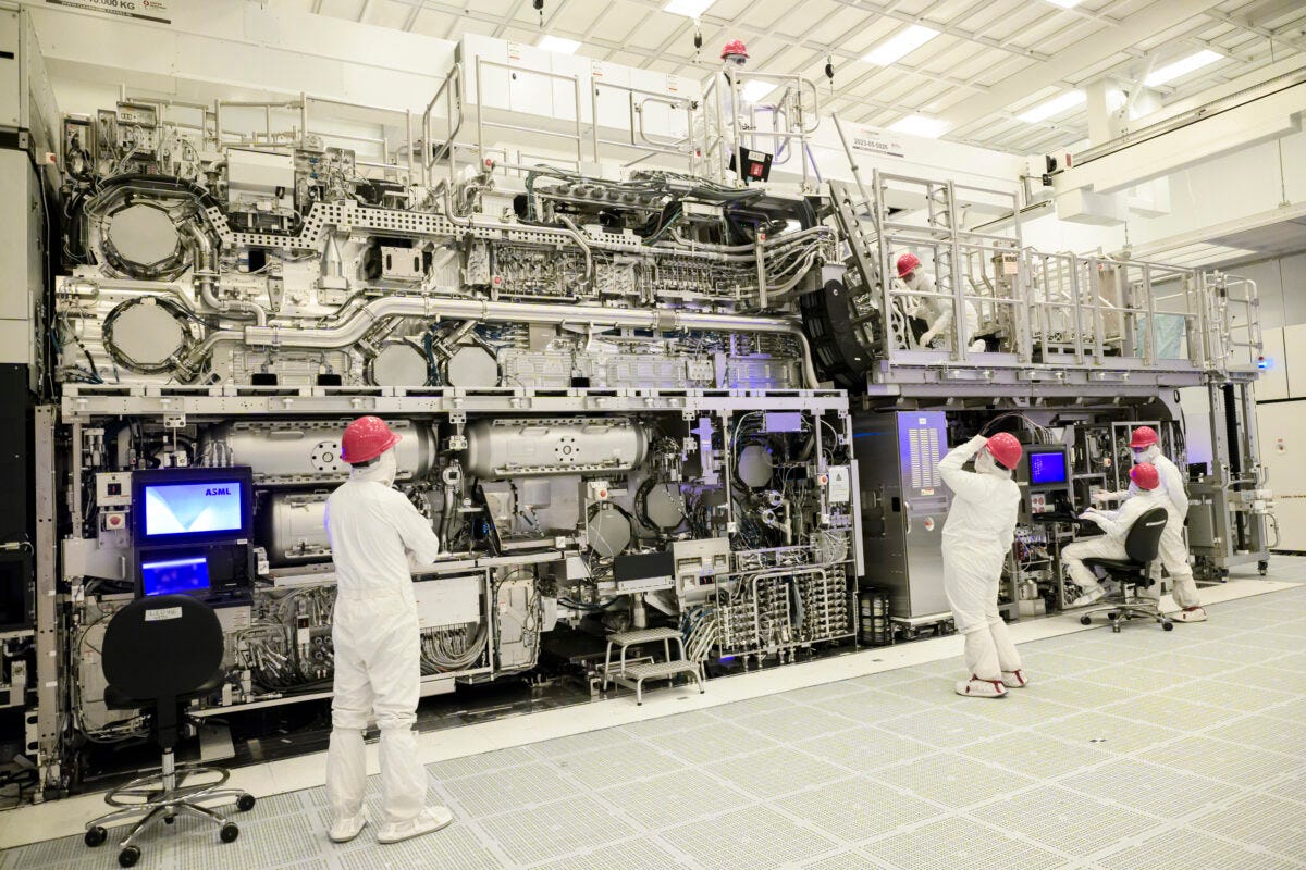 Technicians in white cleanroom suits and red caps work on a large, complex machine with numerous pipes and control panels in an industrial setting. Some use computers, while others inspect the equipment.