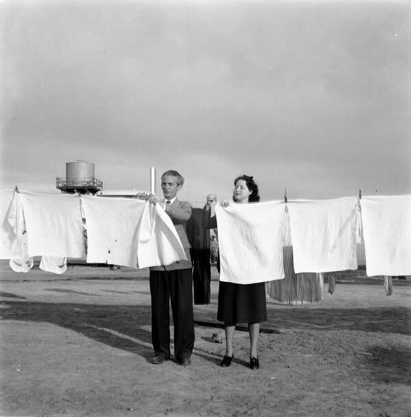A black and white photo shows a man and a woman pinning up white cloths to dry on a clothesline. 