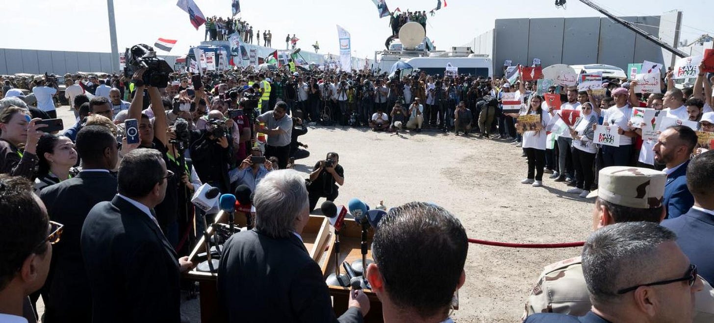 A UN official stands at a podium and speak so press and other civilians bearing signs just outside Gaza.