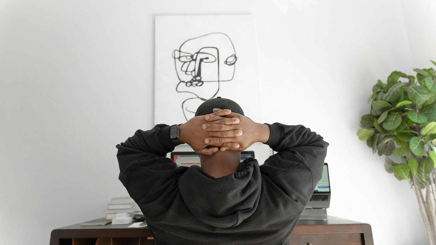 Black creative professional sitting at a minimalist home office desk with hands behind head, viewing laptop screens under abstract art — ideal setup for online art direction course work.