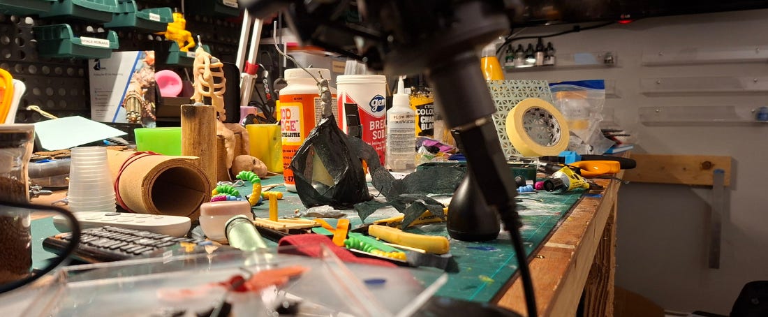 Landscape photo in full colour. It's a photo of my desk, which is very messy. There are a lot of hobby related items on it. Including a tub of mod podge, baking soda, spray paint.