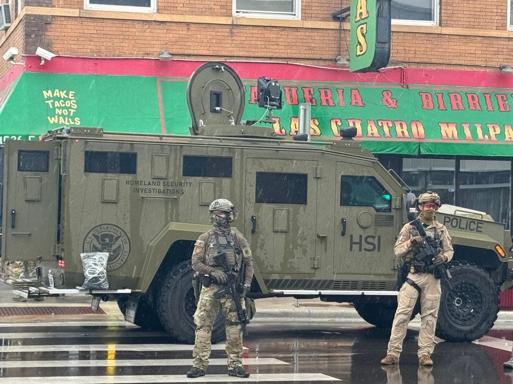 Two ICE agents in full military combat gear (helmets, body armor, camo uniforms, armed with tactical rifles and sidearms) stand in front of a Bearcat armored truck marked with 'Homeland Security Investigations,' a DHS seal, and 'HSI.' An LRAD 'sound cannon' is attached to the roof of the vehicle, and a large clear plastic bag full of dozens of zip-tie handcuffs rests on the rear running board. The battle wagon is parked on the street in front of a taqueria with a festive awning; on the corners of the awning is the slogan 'Make Tacos Not Walls'  (part of its regular decor, not a graffito added for the occupation)