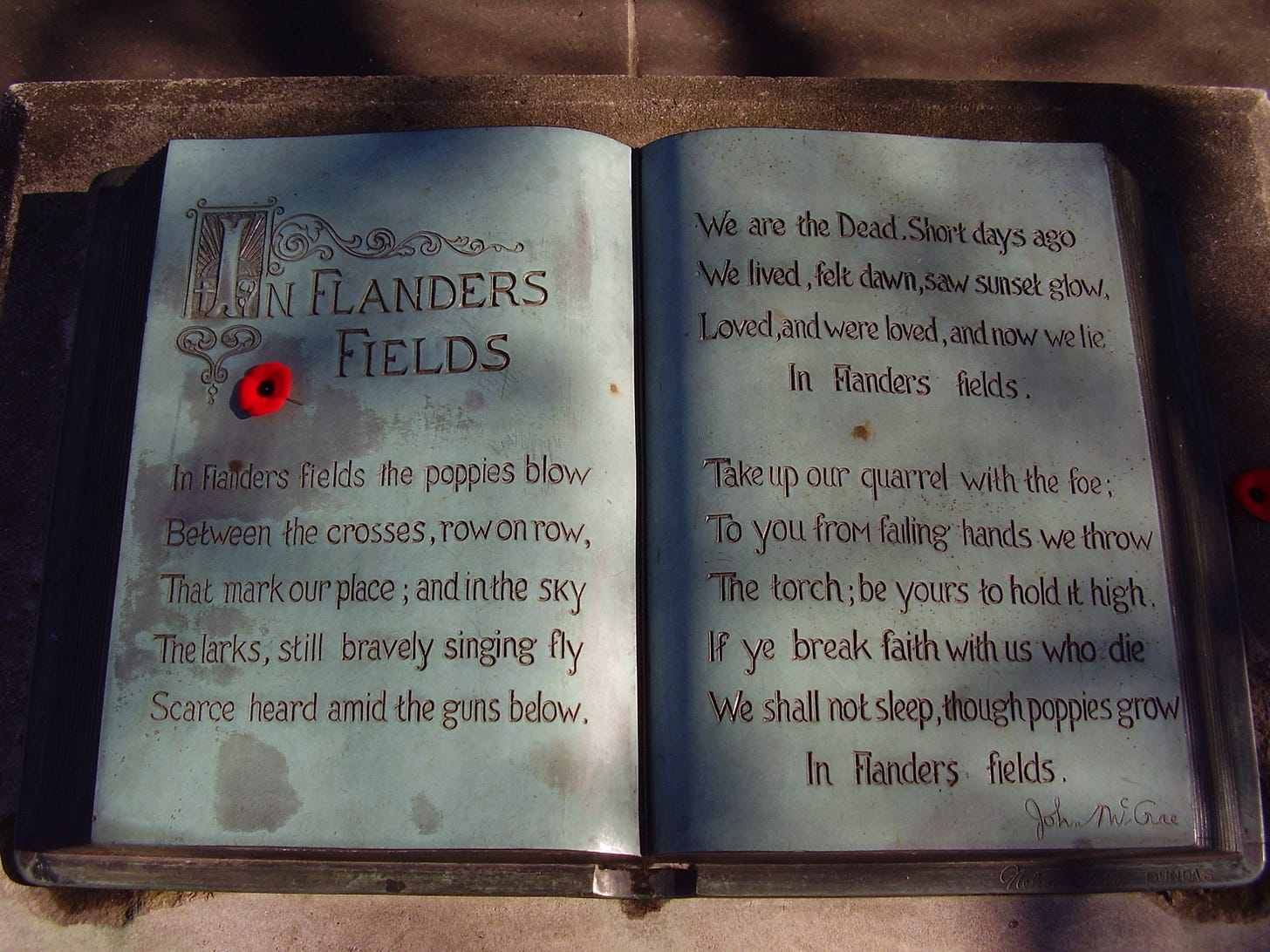 A sculpture in the form of an open book. The text of the poem "In Flanders Fields" is written within and a small red poppy lies on top. A sculpture in the form of an open book. The text of the poem "In Flanders Fields" is written within and a small red poppy lies on top.
