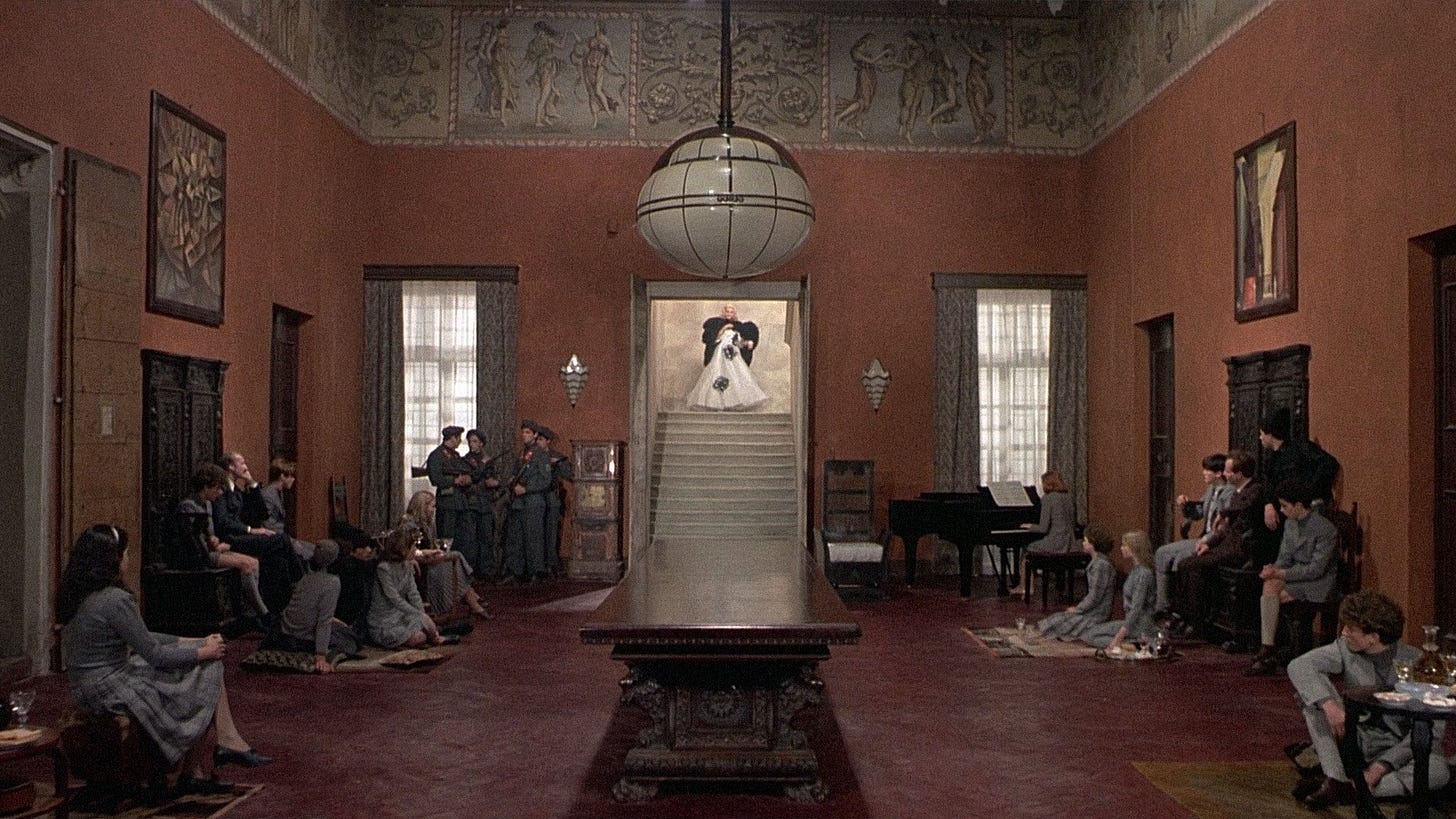 Salò, or the 120 Days of Sodom | Still features a dining hall where students dressed in grey are mostly seated on the floor as they gather to meet a woman in an elegant dress set to descend from a staircase.