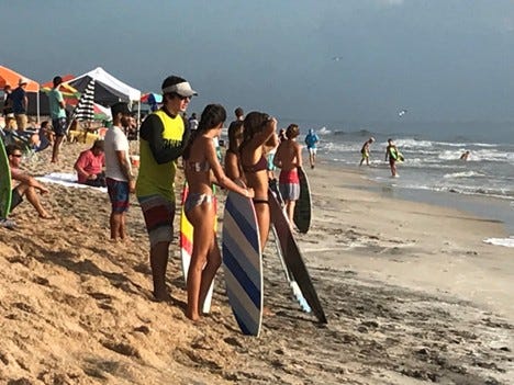 A young man stands on a beach with a group of girls.  They are all holding skimboards.