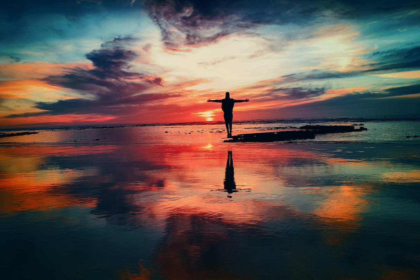 Person on a rock in a reflective body of water with a sunrise or sunset in the background. Person on a rock in a reflective body of water with a sunrise or sunset in the background.
