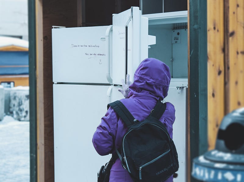 a person wearing a purple coat and backpack opens an outdoor community fridge