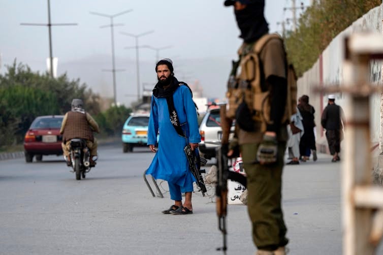 Armed soldiers stand near the entrance of a building as people and traffic can be seen in the background.