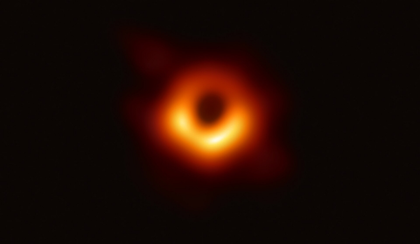 Over a black background of empty space, an orange-red donut of gas lies in the center of the image, with a black circle--the black hole's shadow--in the middle of the donut. Over a black background of empty space, an orange-red donut of gas lies in the center of the image, with a black circle--the black hole's shadow--in the middle of the donut.