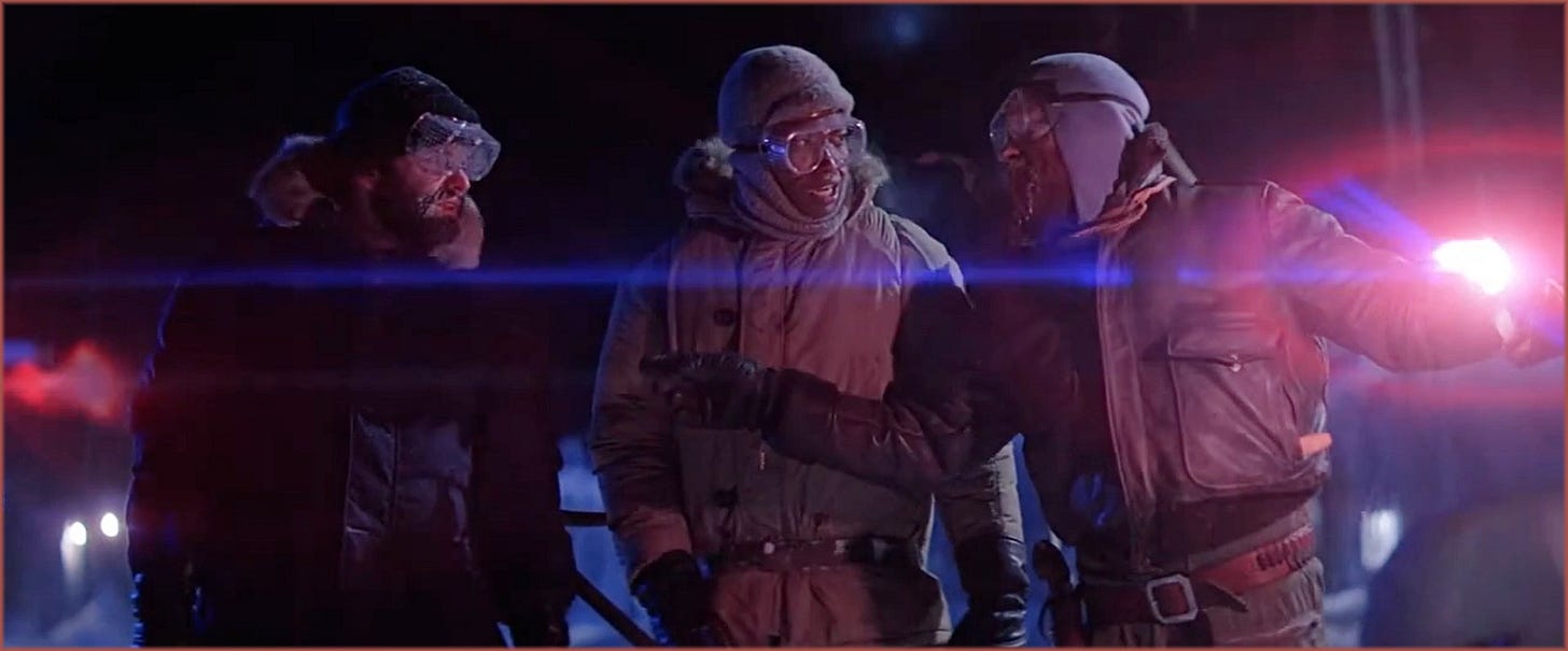Exterior night shot from The Thing (1982): the three men in heavy winter gear stand together in the Antarctic dark, a blue anamorphic lens flare crossing the frame horizontally, magenta road flares burning in the background on the right. Cundey's three-color temperature system — warm amber, cold blue, desperate magenta — visible simultaneously in a single frame.