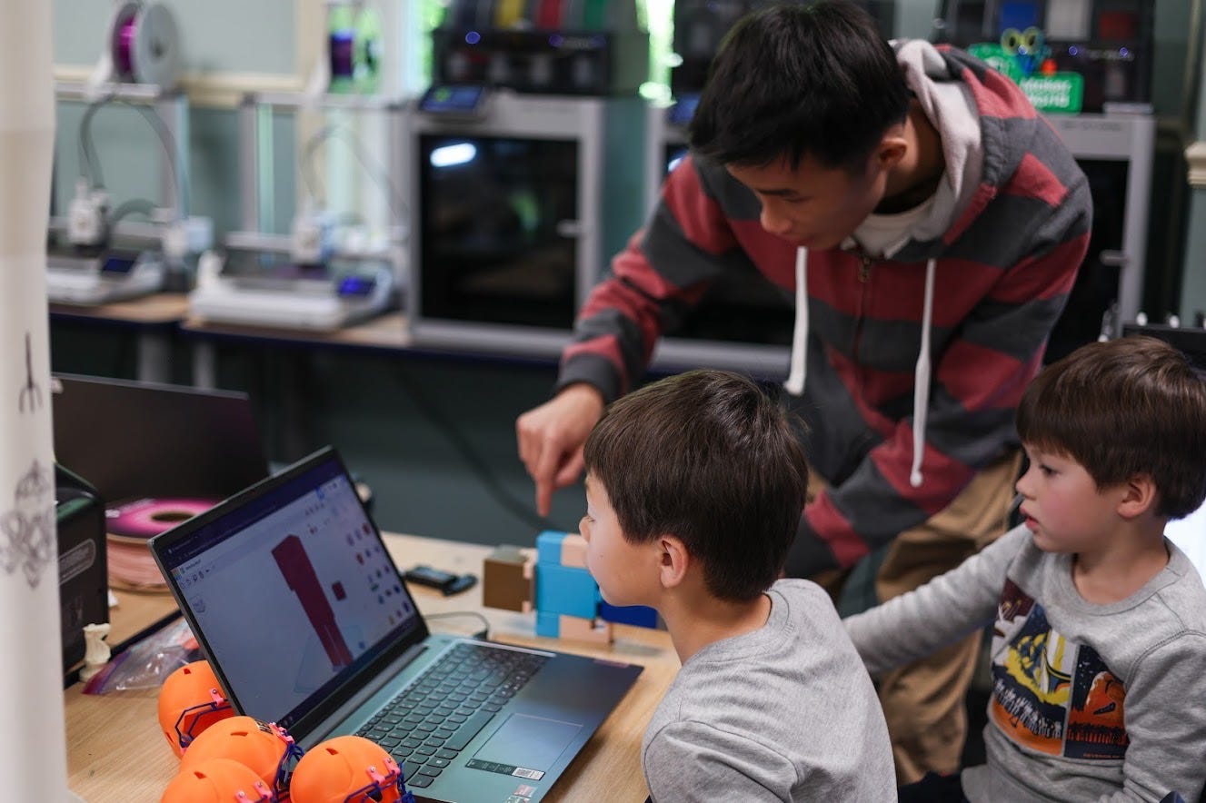 Two young children sit at a table looking at a laptop screen while an adult stands next to them, gesturing at the keyboard. Two young children sit at a table looking at a laptop screen while an adult stands next to them, gesturing at the keyboard.
