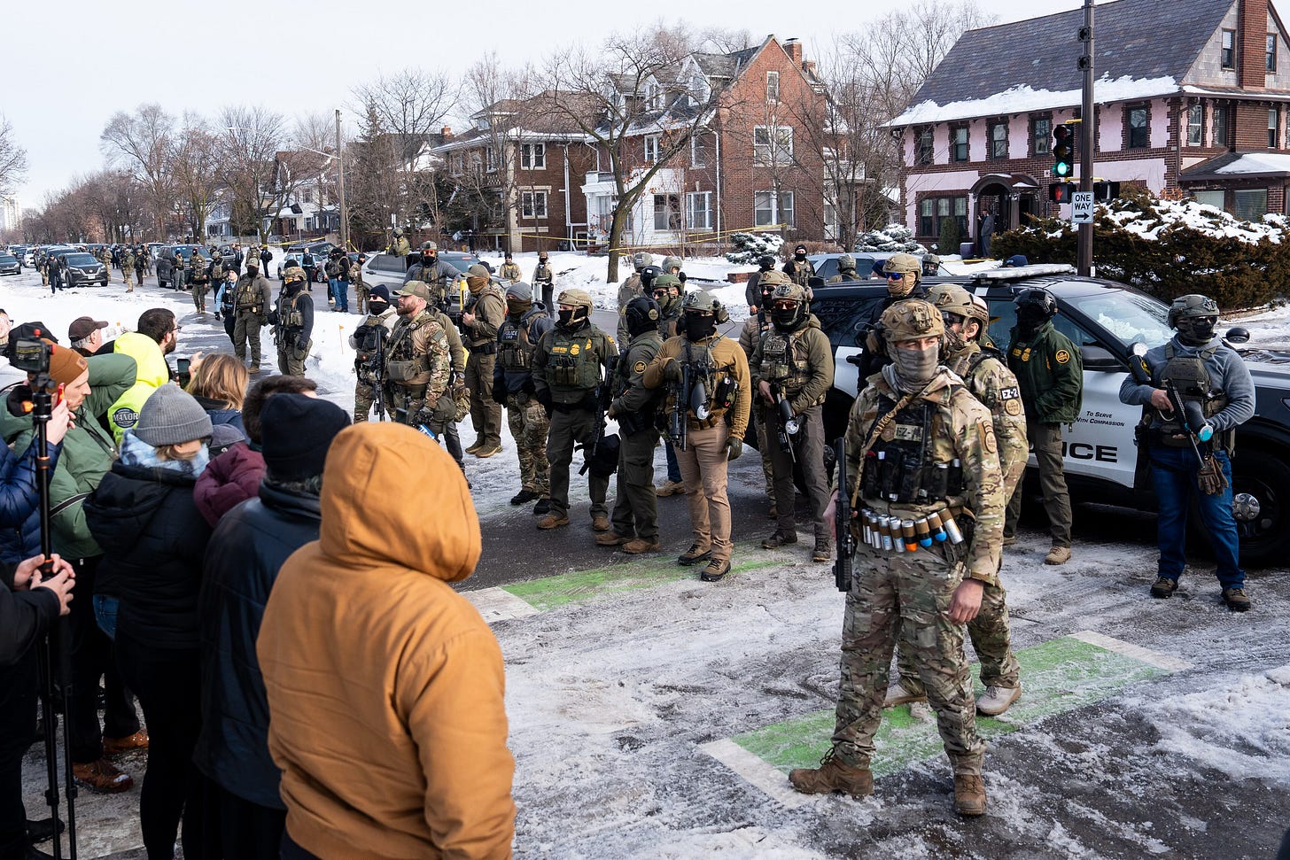 A street-level photograph showing federal ICE agents and local bystanders gathered at an intersection in South Minneapolis following a shooting incident.