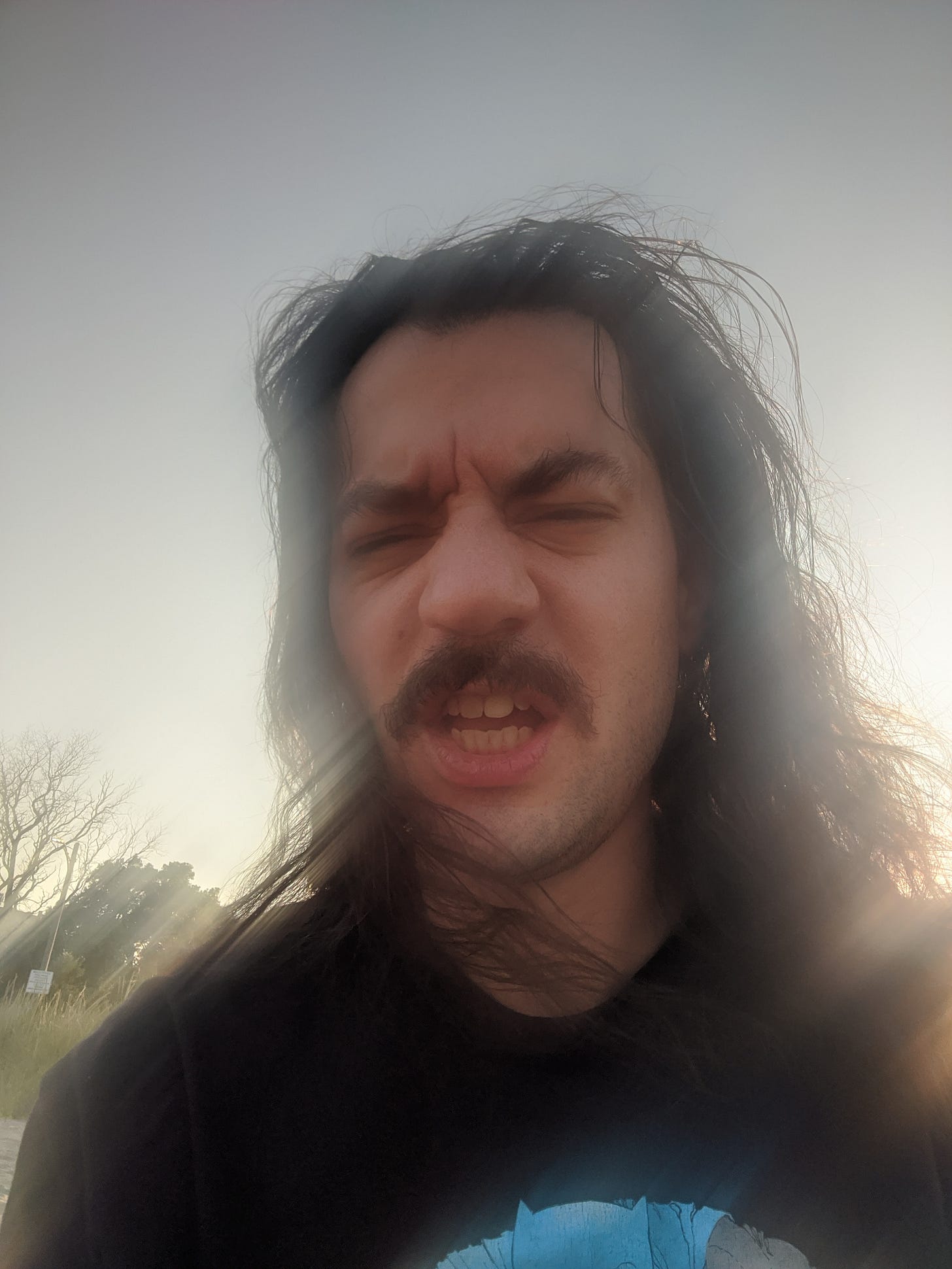 a white man with shoulder-length black hair and mustache is backlit by the fading sun
