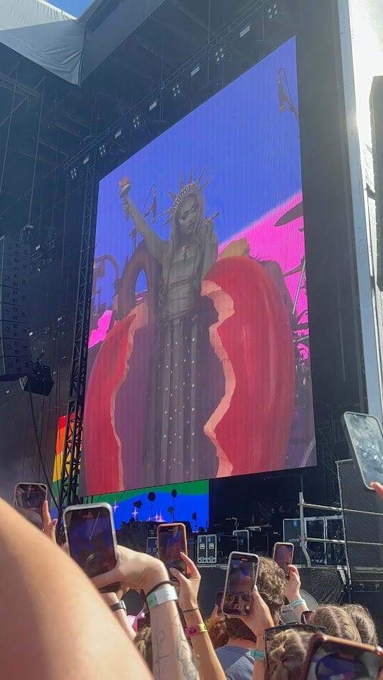 picture of a screen at an outdoor concert. chappell roan, painted green and dressed as the statue of liberty, is standing in a sculpture of a half-eaten apple, in front of a rainbow flag on the screen.