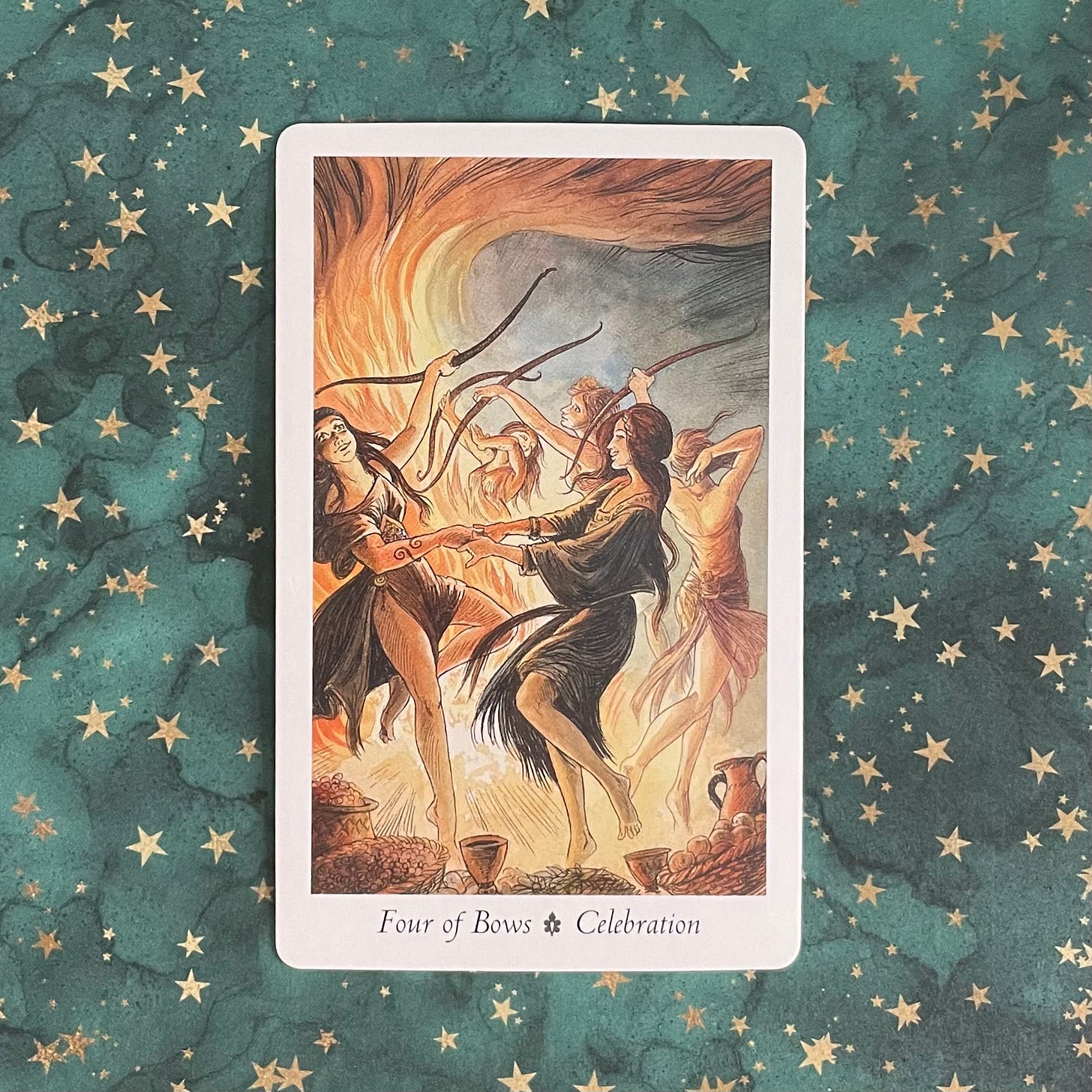 A tarot card from the Wildwood deck sits on a dark green background decorated with golden five pointed starts in various sizes. The card has a white border with a rectangular drawing taking up most of the space inside. In the drawing five women, four of whom are holding unstrung bows above their heads, dance in front of a large bonfire, which is flaming right up to the top of the card. All the women whose faces we can see look happy and relaxed, their bodies seemingly moving to a beat we cannot hear. In the forground are pots and bowls full of food, plus chalices and a jug filled with what I assume to be wine. The words Four of Bows - Celebration are written in black on the white border of the card, directly below the artwork.