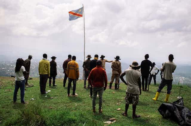 A group of standing men face a speaker standing on a hill overlooking a town below with the Congolese flag fluttering above them A group of standing men face a speaker standing on a hill overlooking a town below with the Congolese flag fluttering above them