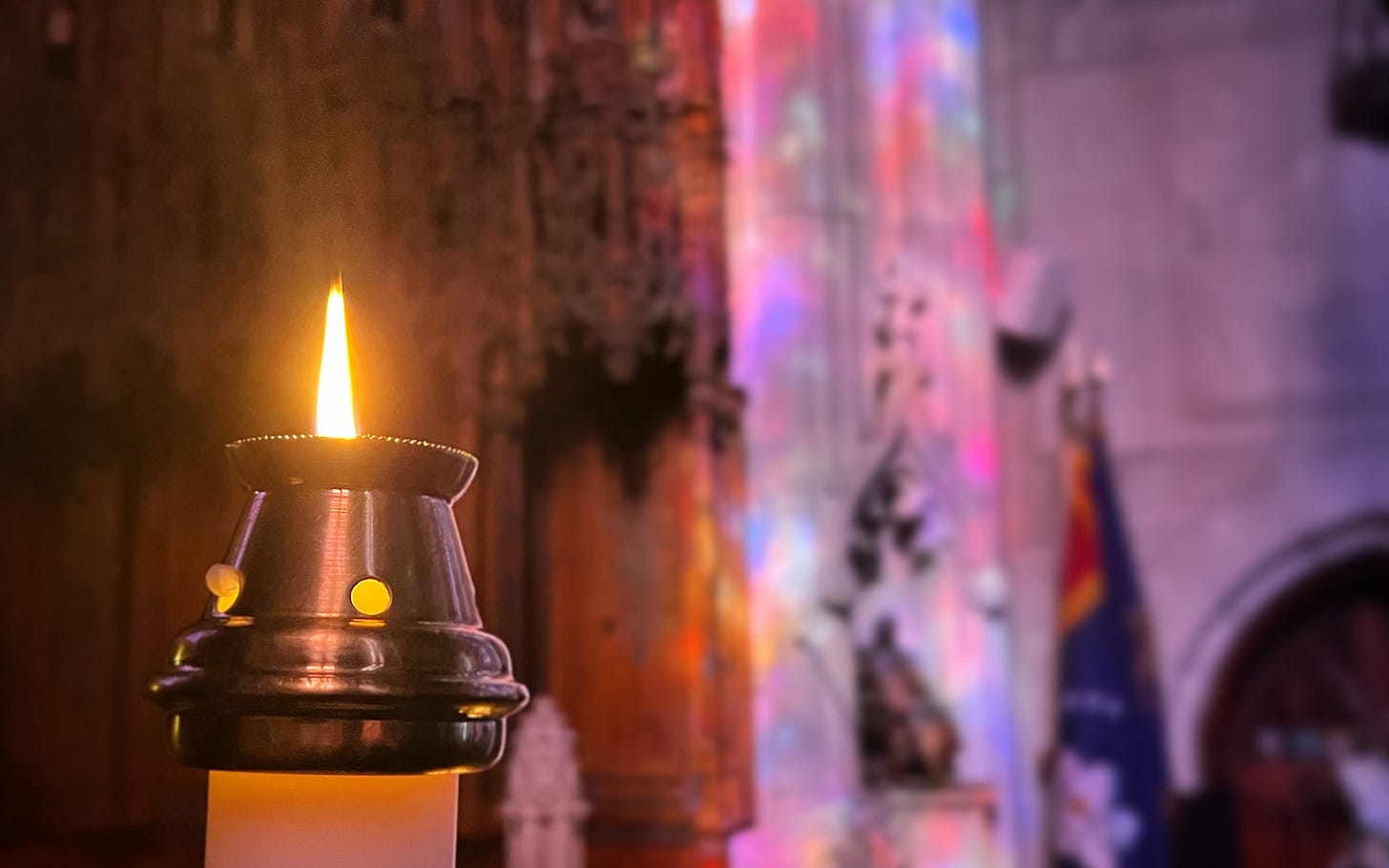 A paschal candle with a brass follower, burning in a church lit only by sunlight coursing through the stained-glass windows.