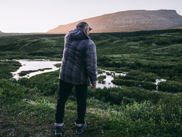 From behind, a man wearing a plaid jacket and dark pants stands on a grassy hill. He gazes across a landscape of green foliage, ponds, and a large hill.