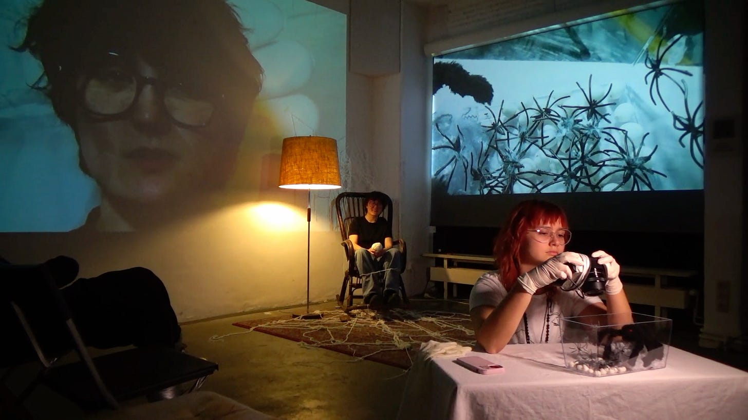 Performance still: A dimly lit performance space with a warm floor lamp. A person with red hair and glasses, wearing gloves, examines a container filled with small objects, while another person sits in a rocking chair in the background. Large video projections of a face and abstract organic shapes fill the walls, contributing to the immersive environment.
