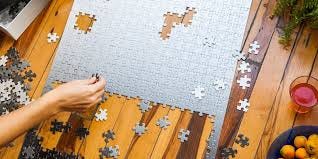 A partially completed white and gray gradient jigsaw puzzle being assembled on a table. A hand, some pieces, and other items are visible on the table.