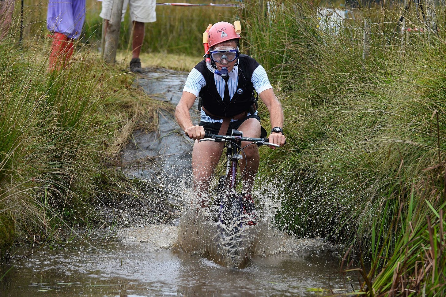 An action photograph of a competitor riding a mountain bike directly through a muddy water trench in a peat bog. The rider is splashing water everywhere and wearing a pink safety helmet, a snorkel, and a diving mask, surrounded by tall, thick marsh grass.