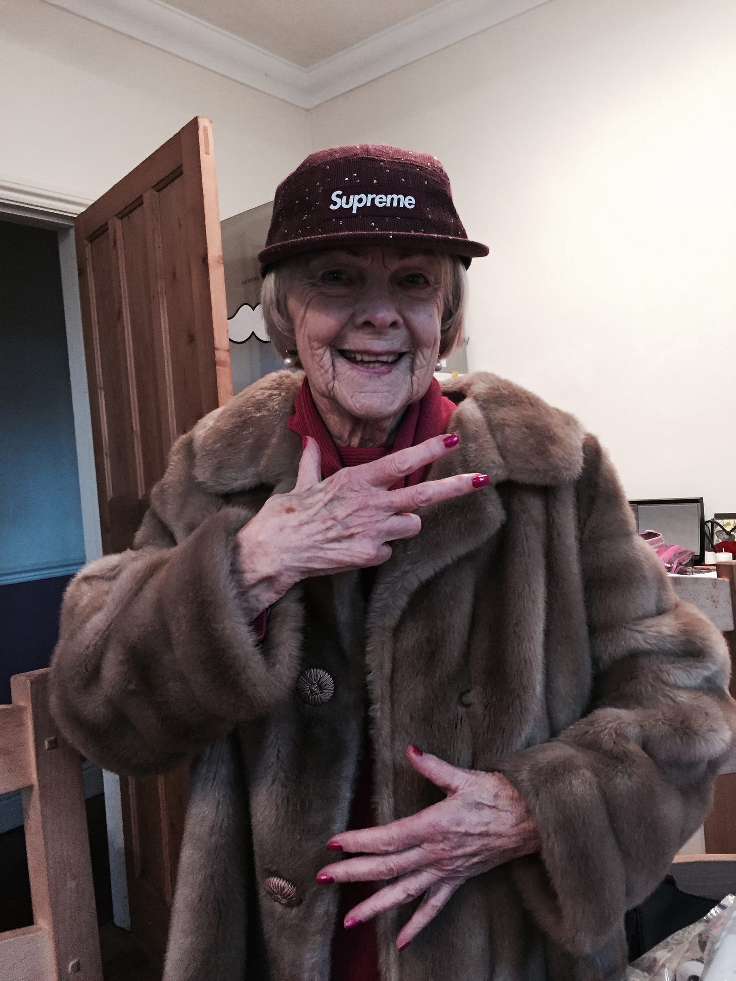 An older lady with pearl earrings, a sparkly Supreme cap, and fur coat posing and smiling for the camera.