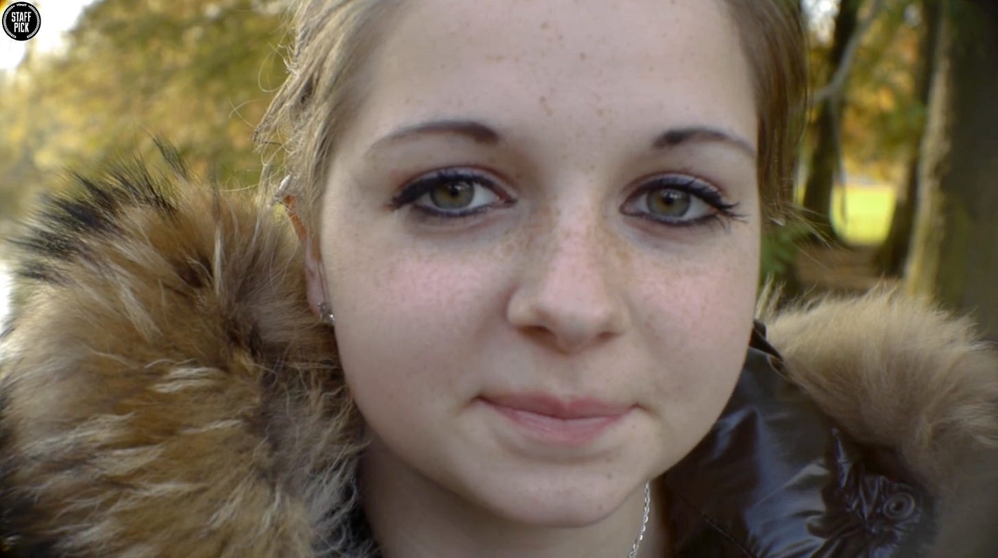 Close-up portrait of a white teenage girl with blonde hair pulled back, wearing a fur-hooded coat and earrings, looking directly at the camera outdoors in fall or winter.
