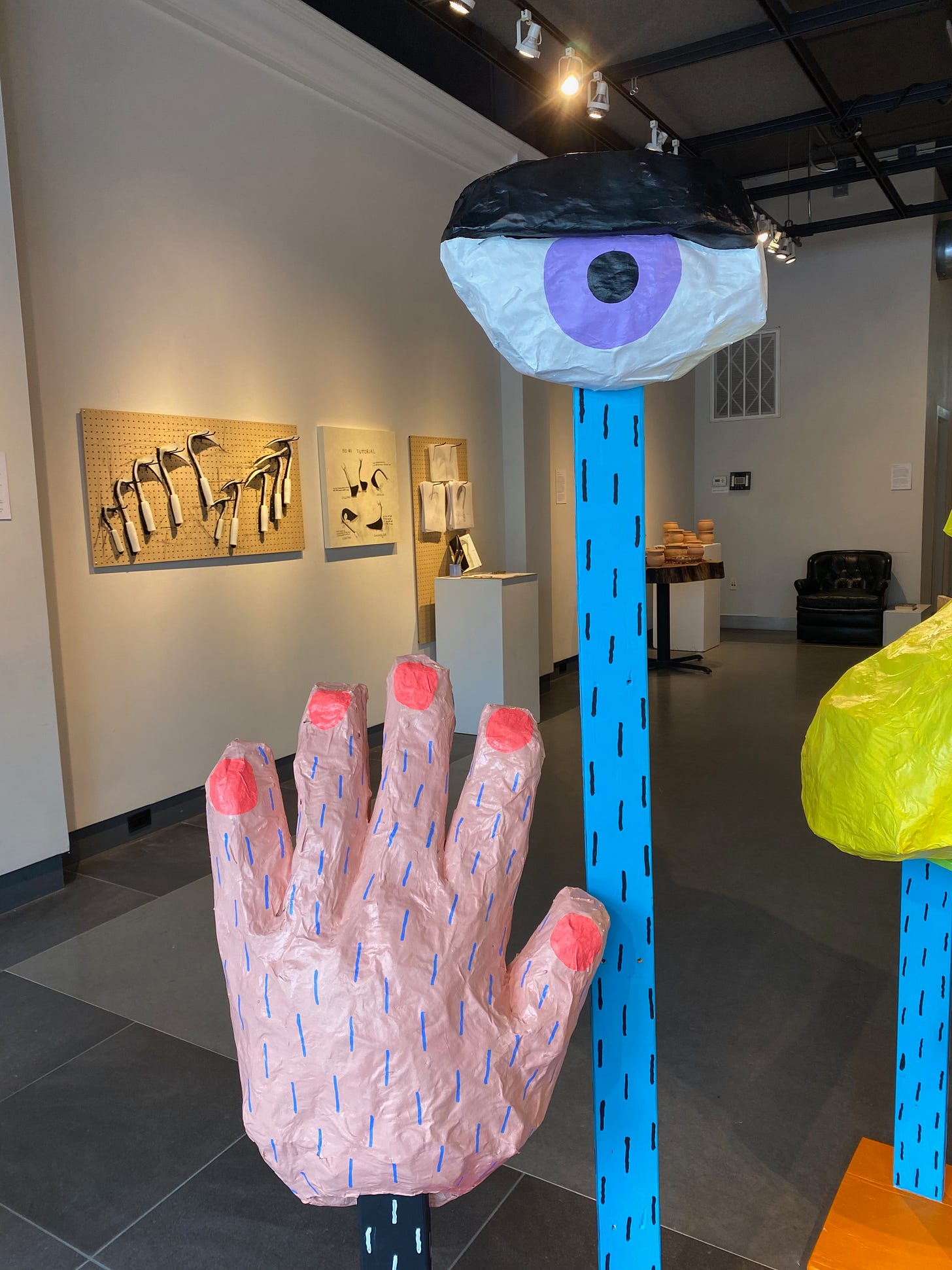 Two huge colorful puppets, one a hand with pink nails, the other an open eye perched on a tall blue stand.