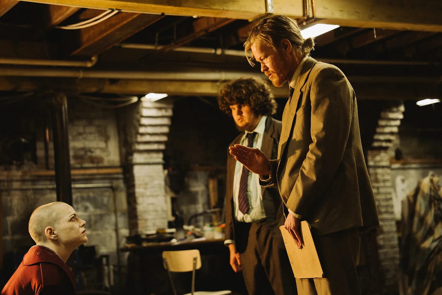 Teddy and Don and Michelle in the basement; Michelle is in the corner of the screen looking up in a red blouse, head shaved; Teddy adn Don are in jacket and tie and lean over her threateningly (with a good bit of hair both). Teddy's holding a folder aginast his legs.