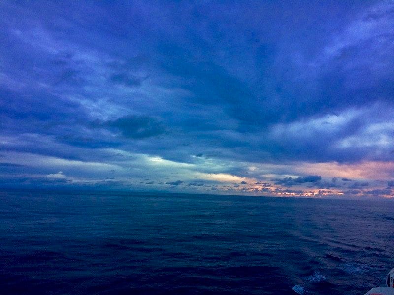 Rainbow colored light sits on the horizon under dark clouds and above a dark sea. Photo by Anna Loscotoff, 2016.