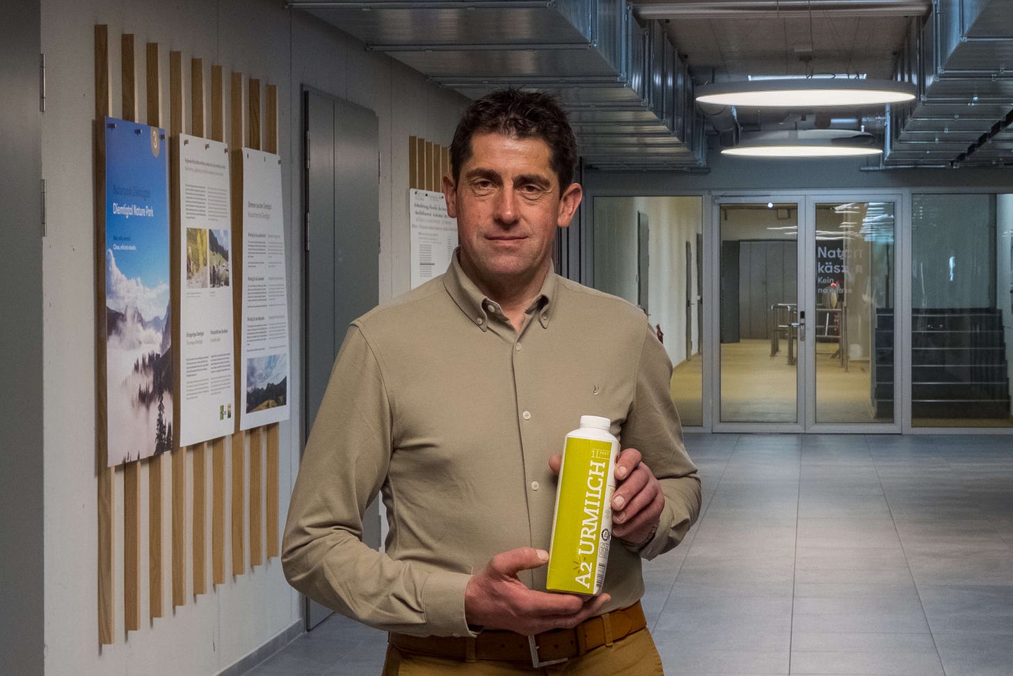 Das Foto zeigt Hansueli Jungen, Landwirt und Vizepräsident von Aaremilch sowie Initiant der Molkerei und Käserei „Simmental Switzerland“. Er steht in einem modernen Flur mit Informationspostern an der Wand und hält eine Packung „A2-Urmilch“ in den Händen. Er trägt ein beigefarbenes Hemd und eine passende Hose, wirkt freundlich und zuversichtlich. Im Hintergrund sind Glastüren und ein hell beleuchteter Korridor zu sehen, was eine professionelle Atmosphäre vermittelt.