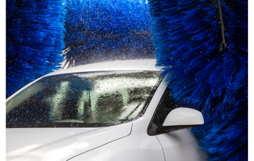 A white car sits inside a carwash, surrounded by bright blue, spinning brushes and droplets of water.
