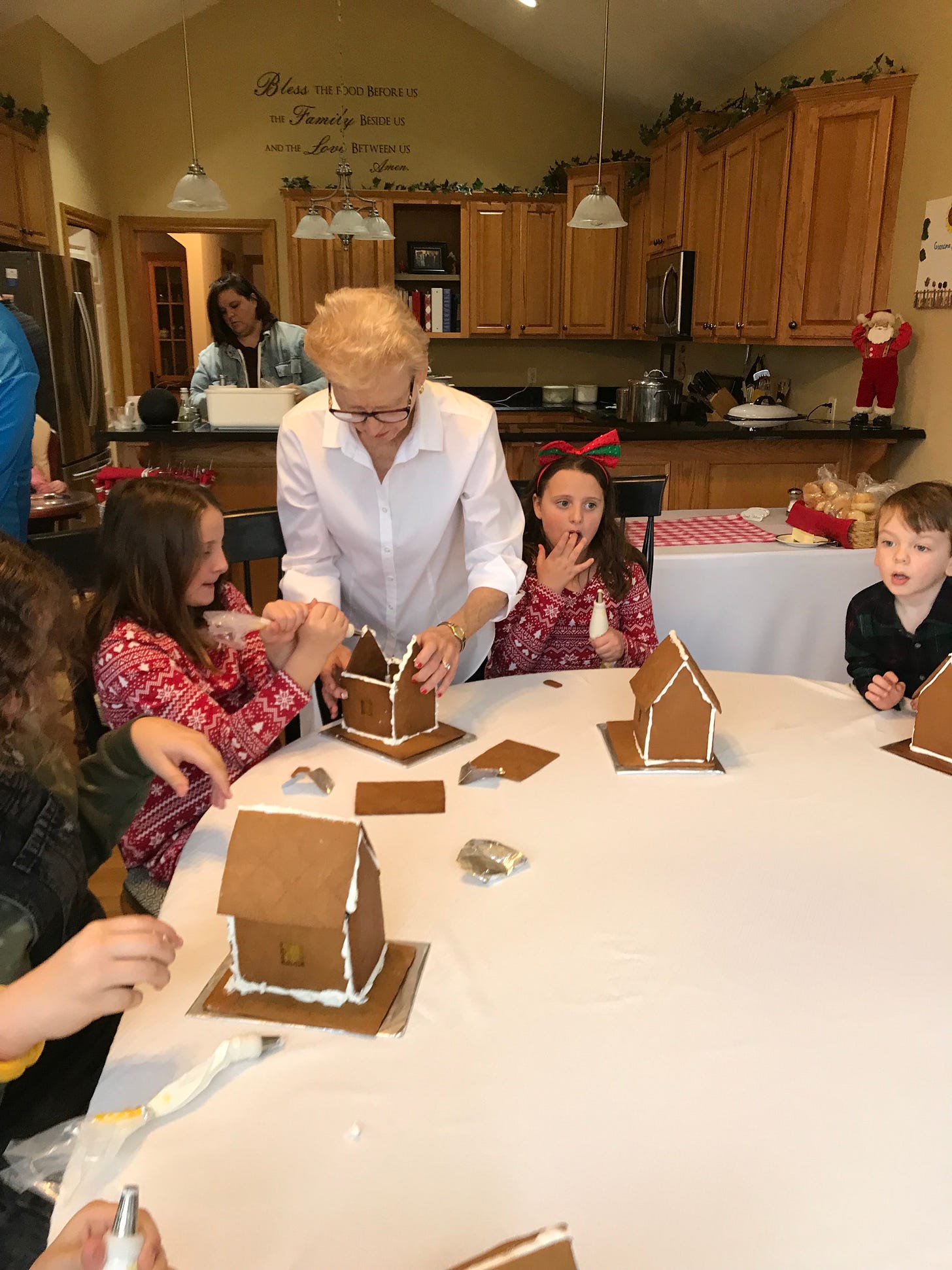 M.A. Hastings in white blouse holding sides of a gingerbread house being constructed with gingerbread cookie panels and white icing by M.A.’s identical twin grand nieces.  The 11-year-old Grand nieces are dressed alike in red and white long-sleeved dresses with large red bows in their hair.  One is holding an icing tube to the gingerbread house in front of her, while the twin on the other side of M.A. at the large table is licking icing off her fingers from her putting together her house.