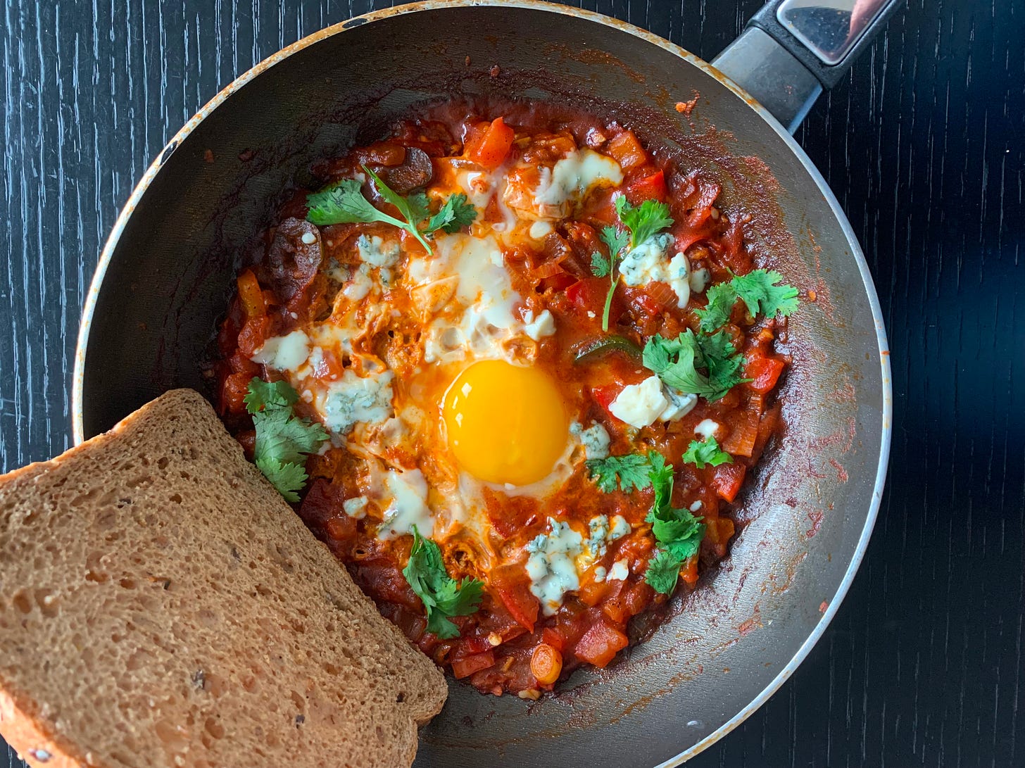 A skillet of shakshuka topped with crumbled blue cheese and fresh cilantro leaves, paired with a slice of dark rye bread. The combination of bold flavors and textures tells a story of creative spins on the classic dish. Made by Liat Portal, showcasing her culinary artistry on The Liat Show and Foodie Disorder.