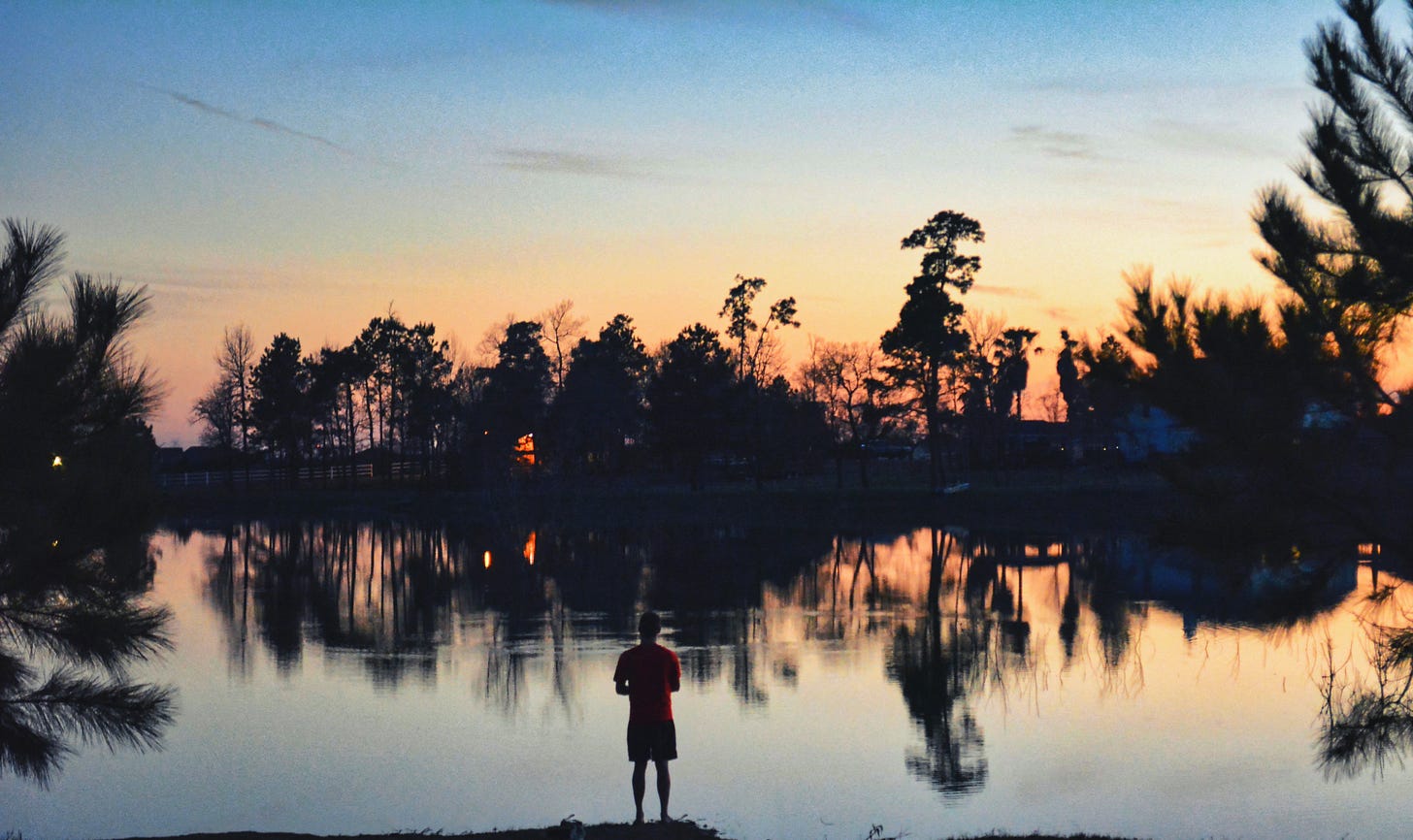 Sunset on the lake with the shore and sky refelected in the water and a fisherman in the center of the foreground facing the lake