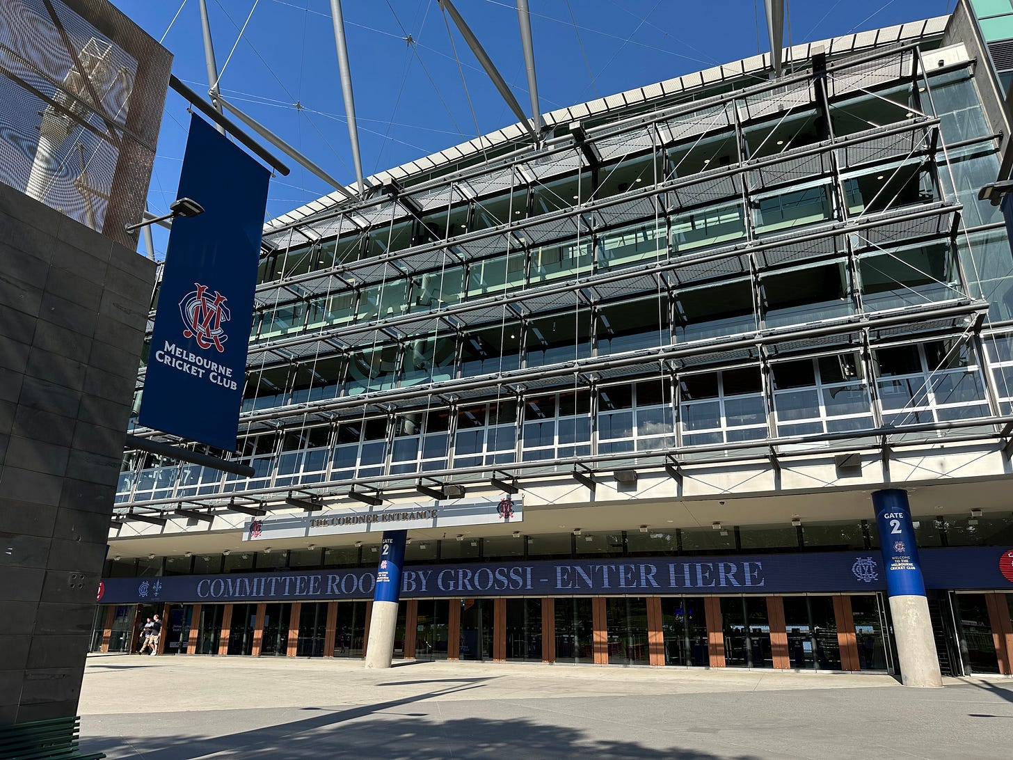 View of the Melbourne Cricket Ground (MCG) exterior on a sunny day, showing the Melbourne Cricket Club banner and the Members’ Entrance with signage for the Committee Room. The stadium’s glass façade and structural framework are clearly visible.