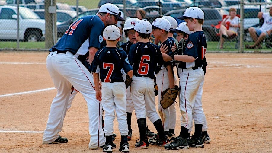 A youth baseball coach kneels and leans into a small huddle of young players in navy jerseys and white pants on a dirt infield, offering guidance during a game while spectators and cars are visible beyond the outfield fence. A youth baseball coach kneels and leans into a small huddle of young players in navy jerseys and white pants on a dirt infield, offering guidance during a game while spectators and cars are visible beyond the outfield fence.