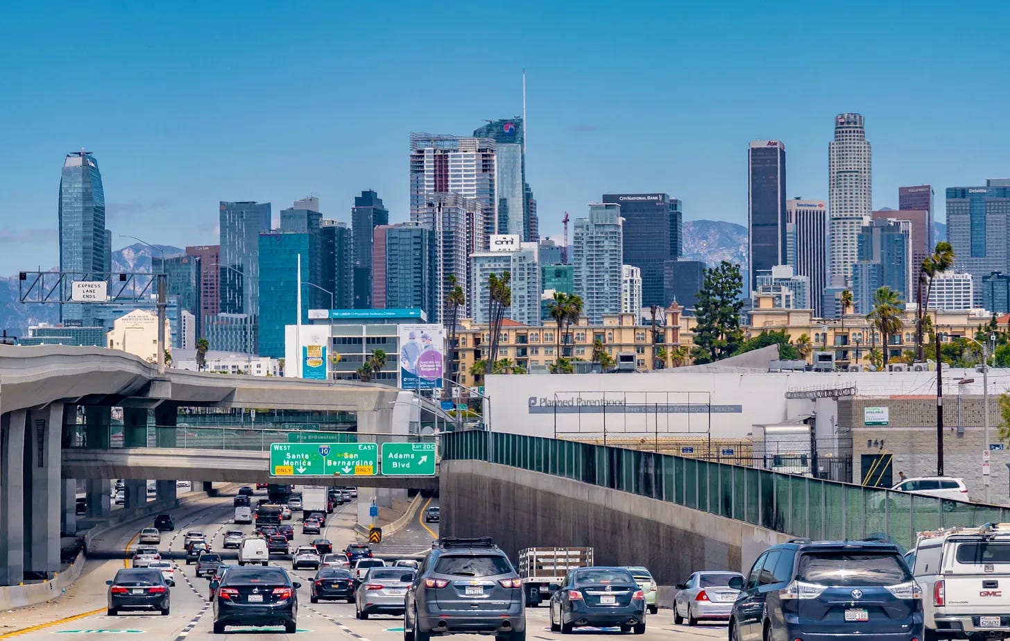Heavy vehicle traffic on a multi-lane Los Angeles highway leading toward the high-rise downtown skyline under a clear sky.